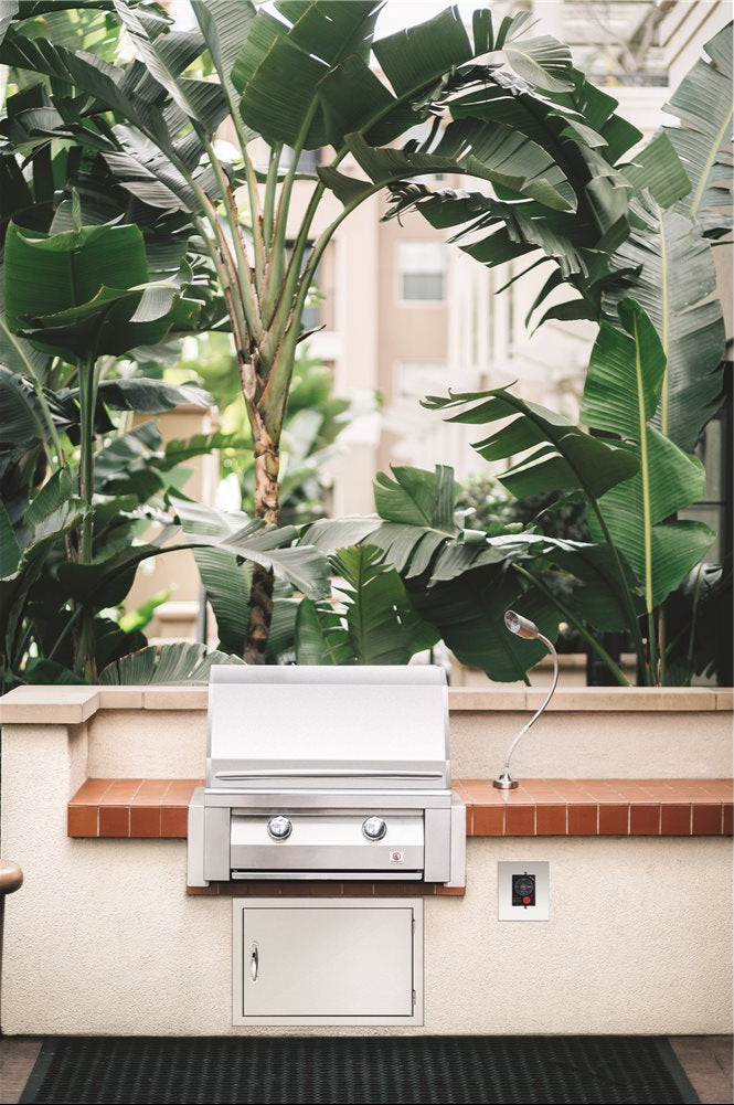 27-inch built-in drop-in stainless steel two-burner gas grill with lower access door and gooseneck light in tile-capped outdoor kitchen island, tropical banana-leaf backdrop