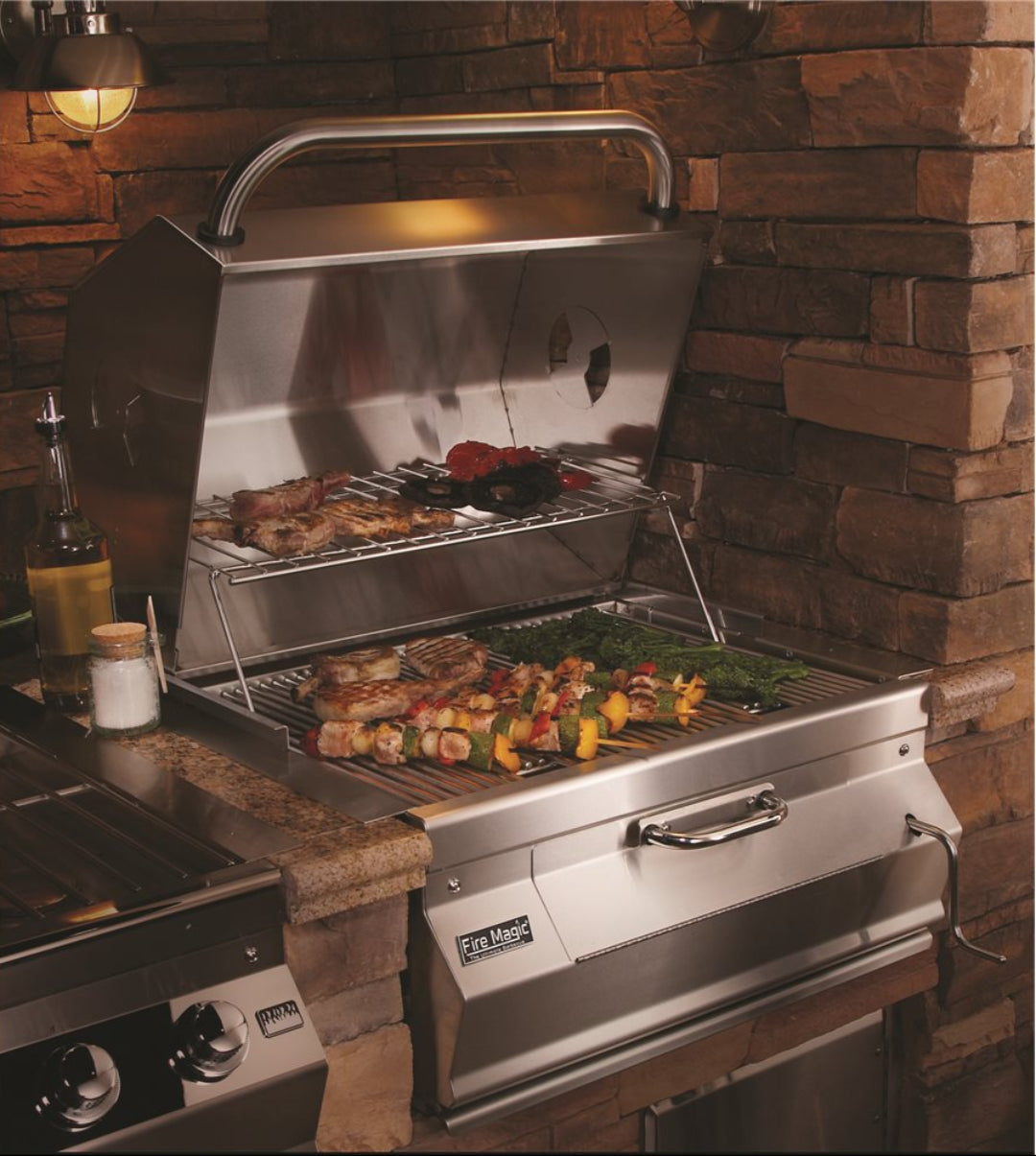Fire Magic stainless steel built-in gas grill for outdoor kitchen island with warming rack, cooking skewers, steaks and vegetables in stone surround