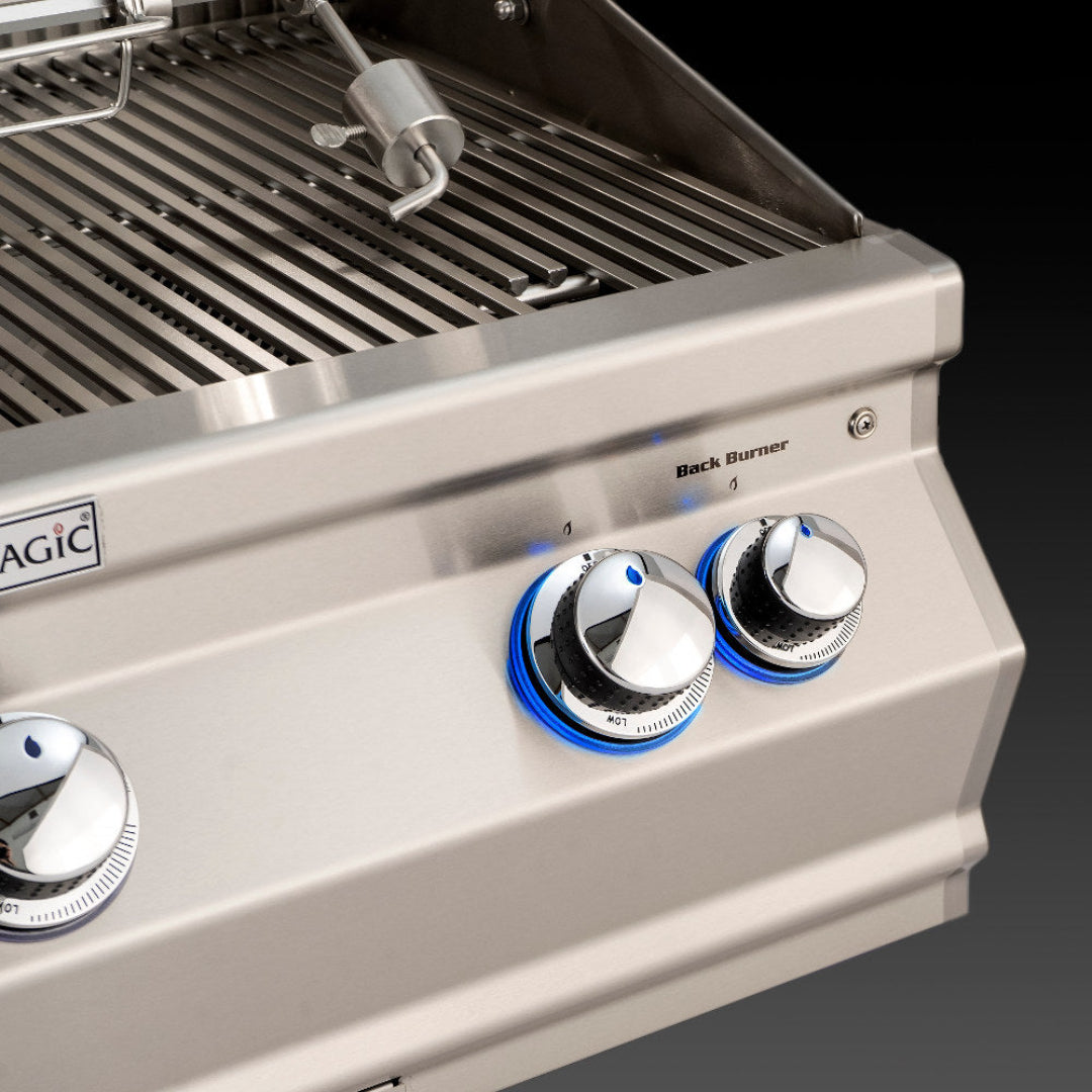 Fire Magic stainless steel built-in gas grill control panel close-up with blue LED illuminated chrome knobs, "Back Burner" label, stainless sear grates and rotisserie spit
