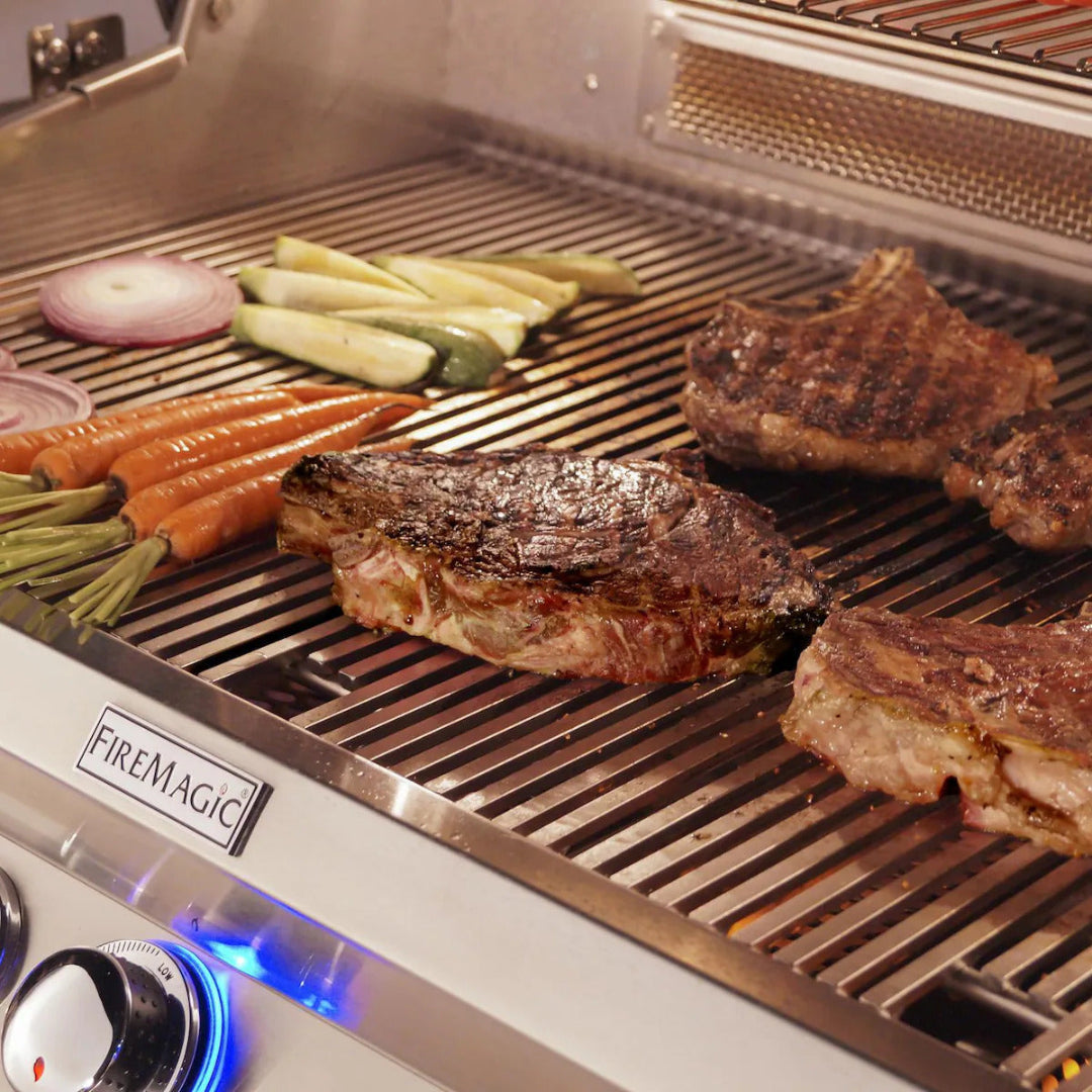 FireMagic A660I stainless steel built-in gas grill searing ribeye steaks and grilled carrots, zucchini and red onion on heavy-duty stainless grates with blue LED control knob for outdoor kitchen