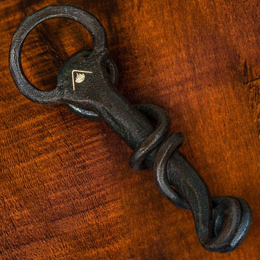 A black Cowboy Cauldron bottle opener with a spiral design and a leather lanyard, displayed on a wooden surface.
