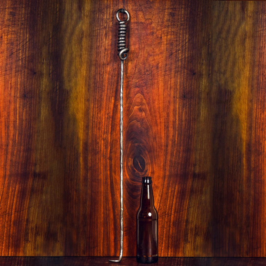 Vine and Flame hand-forged Cowboy Cauldron Pigtail — long wrought-iron hanging cauldron hook with spiral coil handle and L-shaped foot for campfire cooking, shown with beer bottle for scale