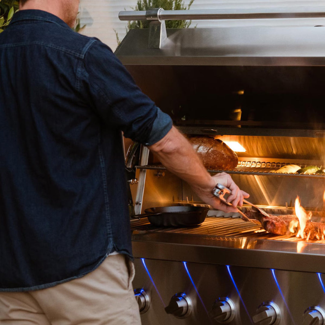 Man grilling steak over open flames inside American Made Grills Encore 54-inch hybrid grill with food on top rack and blue-lit knobs