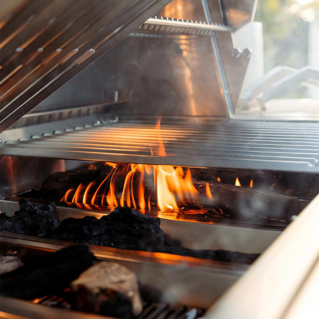 Encore 36-inch hybrid grill with flames beneath stainless steel grates and charcoal visible in firebox