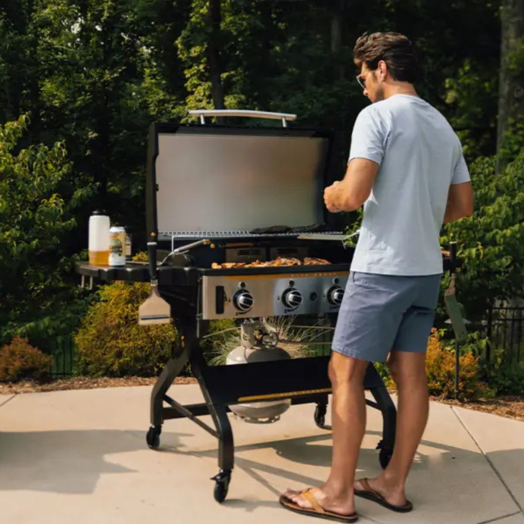 HALO Elite 4B Griddle HZ-1001-XNA outdoor 4-burner flat-top griddle on wheeled cart with open hood and warming rack, food cooking on griddle, propane tank, man grilling.