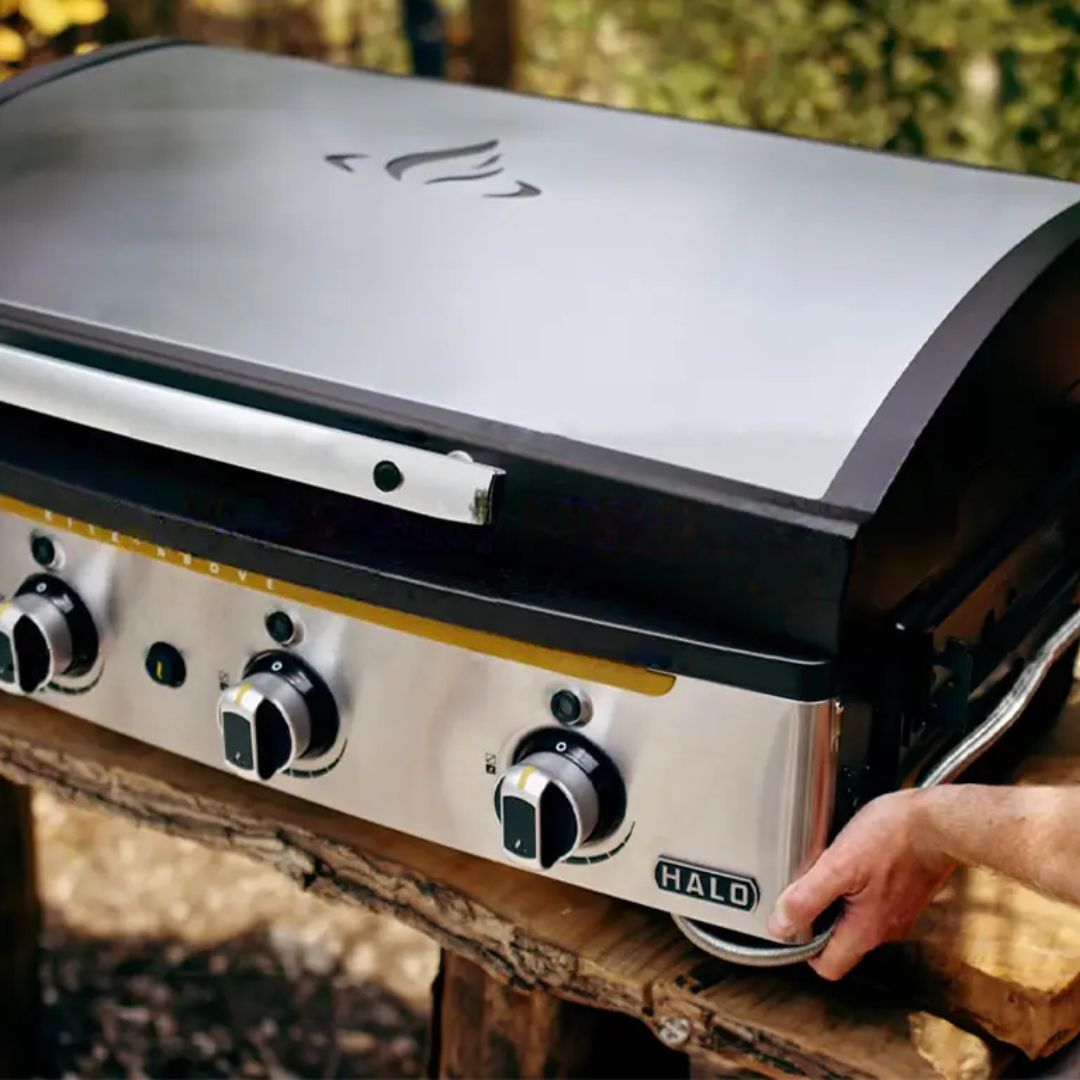 HALO Elite 4B Griddle HZ-1001-XNA stainless-steel 4-burner flat-top grill with closed hood, front control knobs and HALO badge on a wooden outdoor table with a hand at the side.