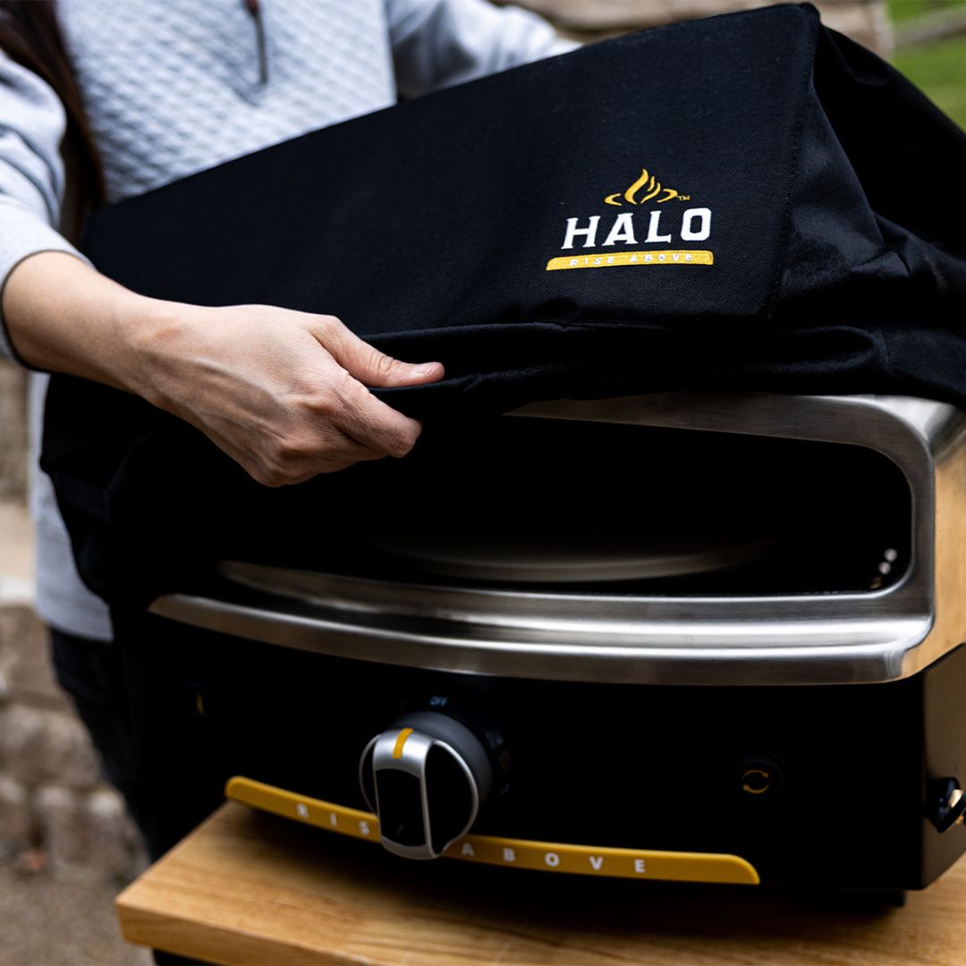 Black HALO Versa 16 pizza oven cover (HZ-5004) with embroidered HALO logo being placed over stainless-steel outdoor pizza oven
