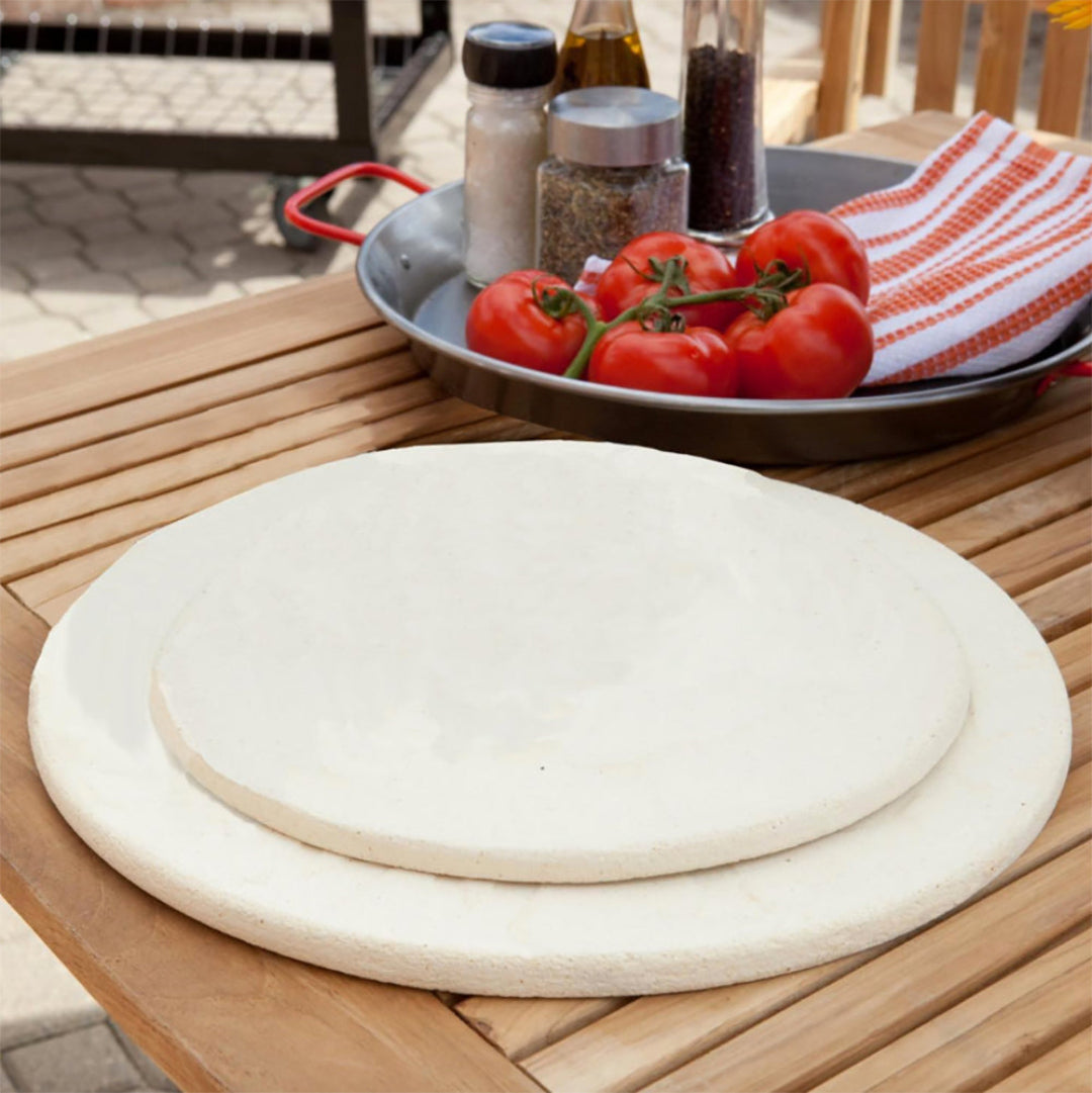 Primo 16-inch natural finish ceramic baking stone on wooden table with fresh tomatoes and spices — ceramic pizza stone for Primo kamado and outdoor grills, heat-retaining baking stone for pizza and artisan bread