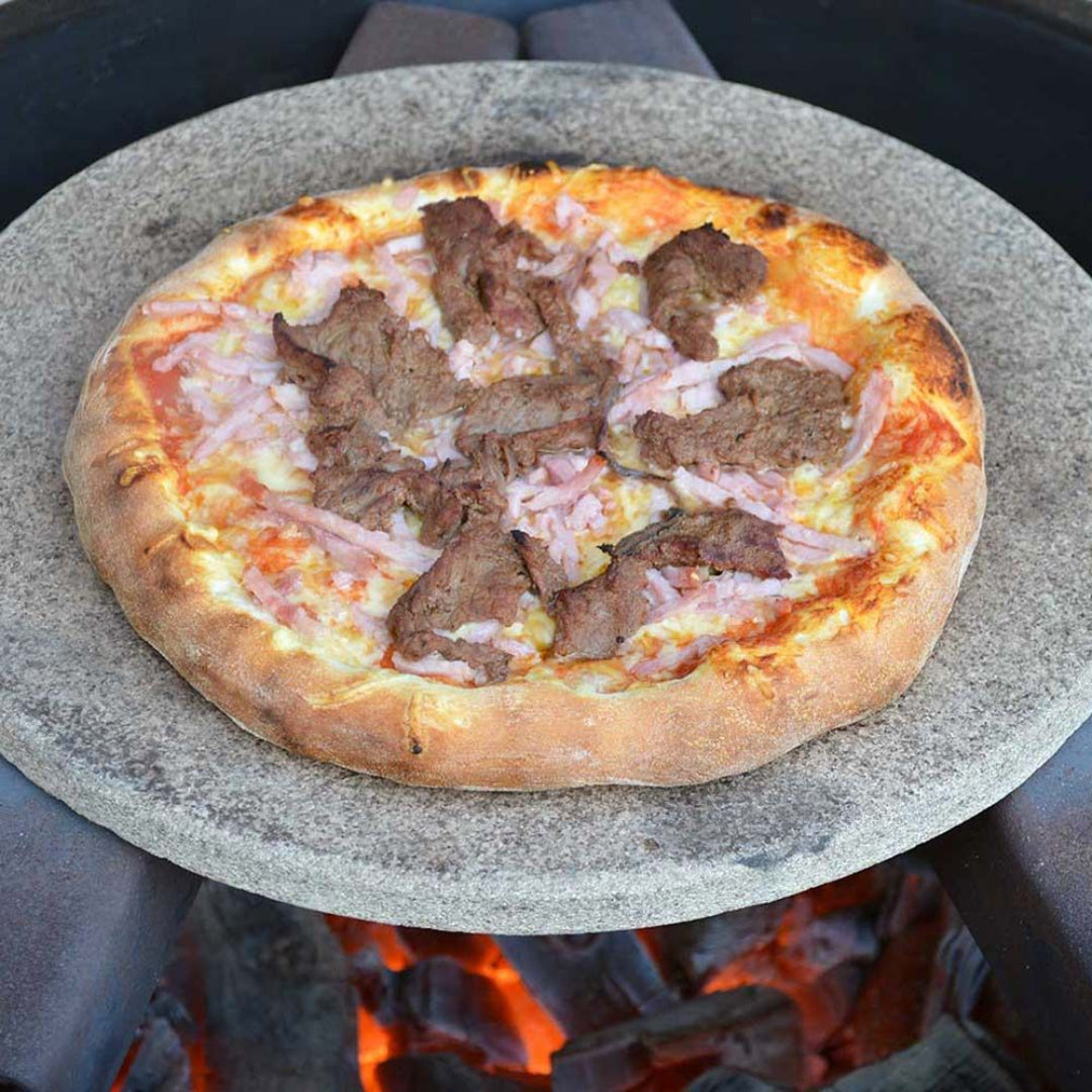 Primo 16-inch round Fredstone pizza baking stone on kamado grill with cooked meat-topped pizza over glowing charcoal