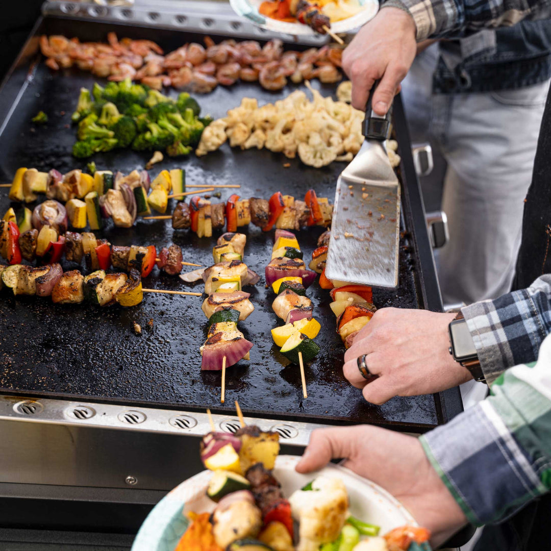 Vine & Flame 36-inch Patio Pro outdoor flat-top griddle cooking meat and vegetable skewers with spatula on seasoned surface