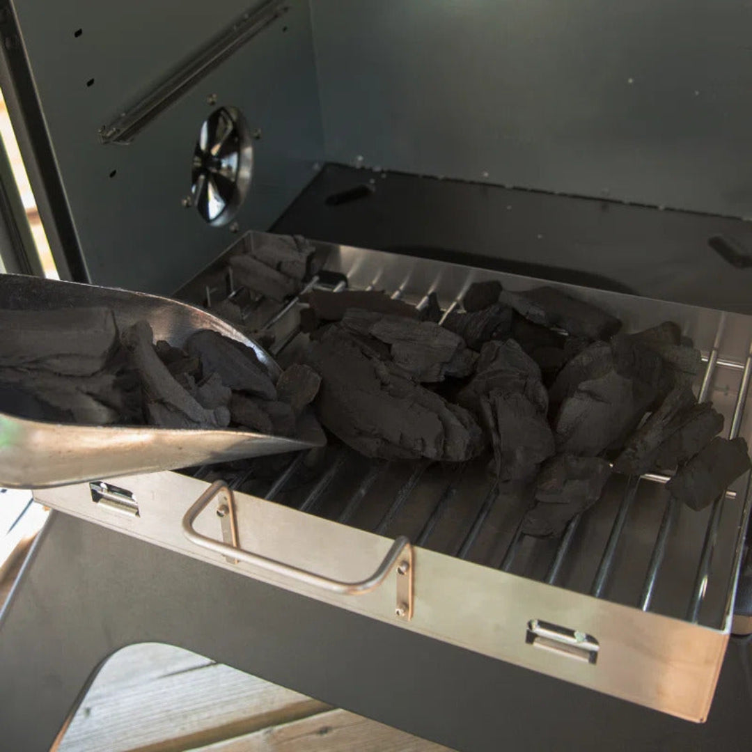Vine & Flame NG5 stainless steel charcoal tray being loaded with natural lump charcoal using metal scoop in portable charcoal grill firebox