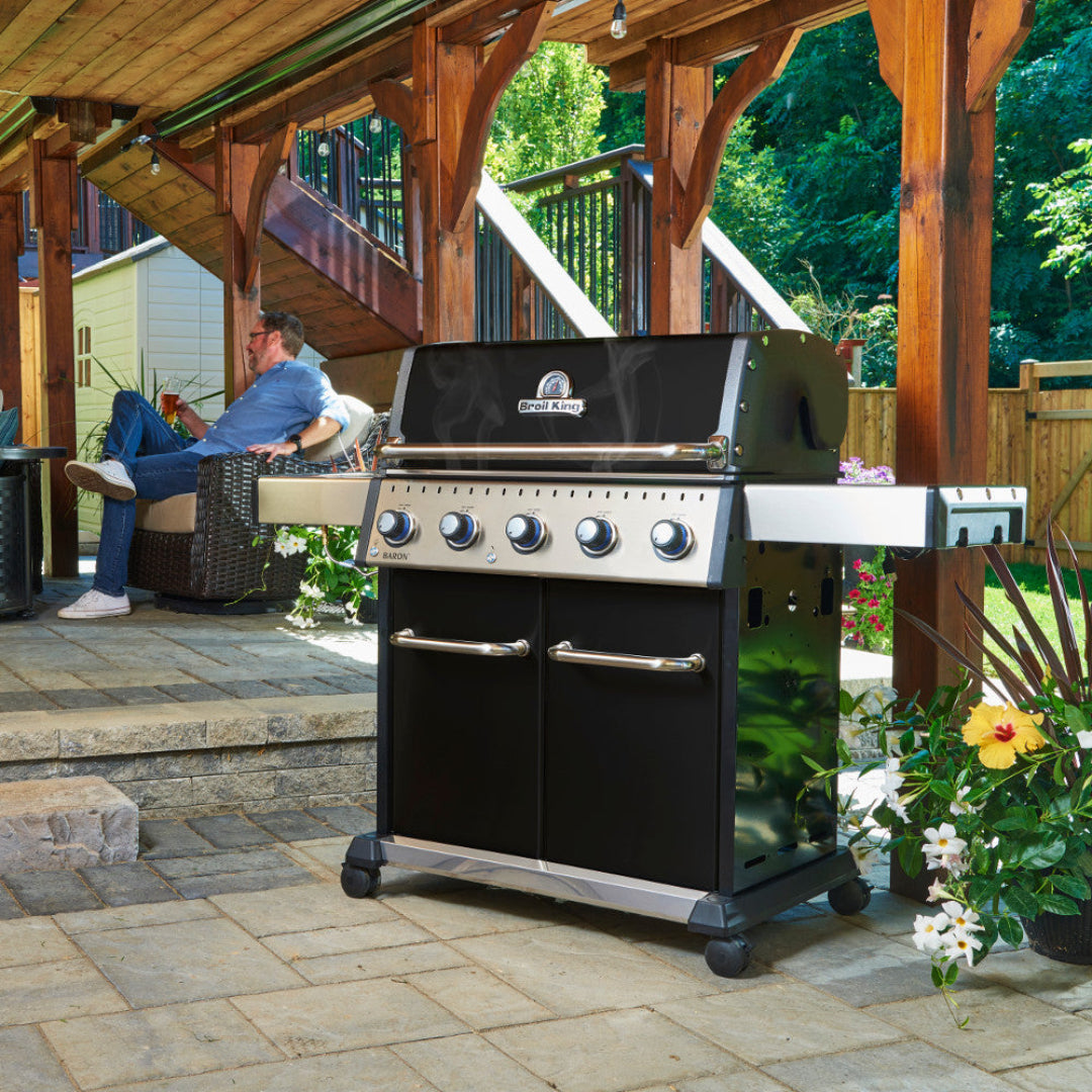 Outdoor grill, Broil King, Baron Series, freestanding propane gas grill on patio with smoke rising and man relaxing in background