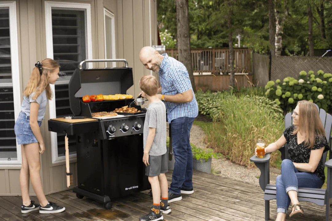 Family grilling on Broil King Crown Series propane gas grill on backyard deck with open lid and assorted food grilling
