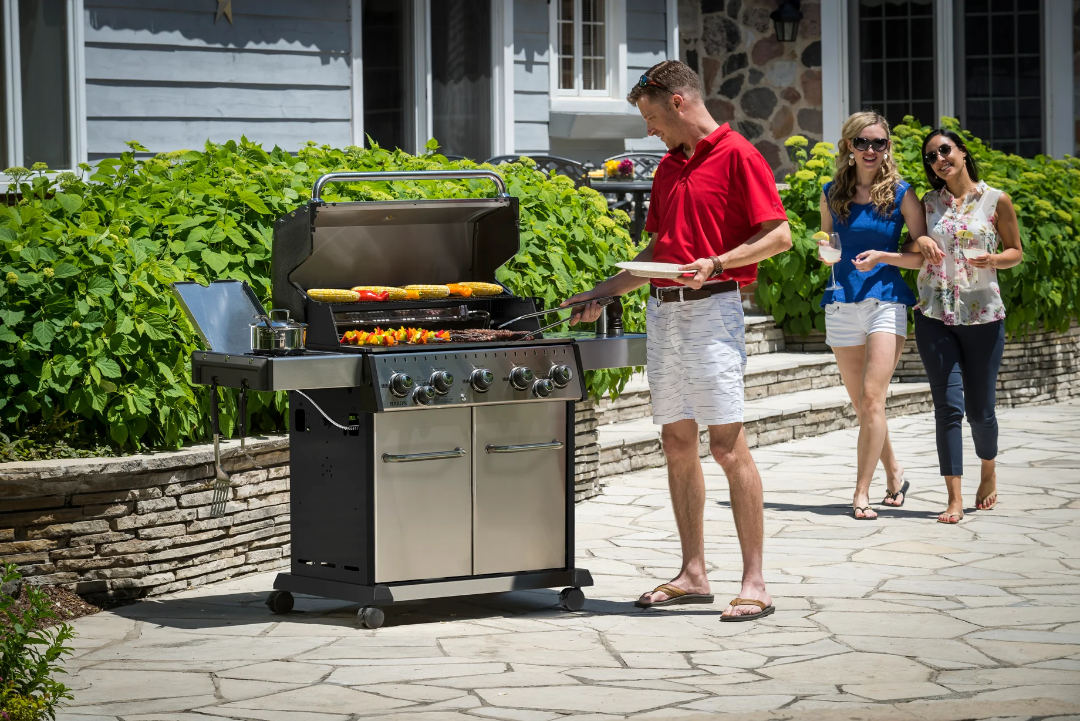 Broil King Regal S 590 Pro 5-burner stainless steel propane gas grill with side burner and double-door cabinet on backyard stone patio, man grilling corn and vegetable skewers