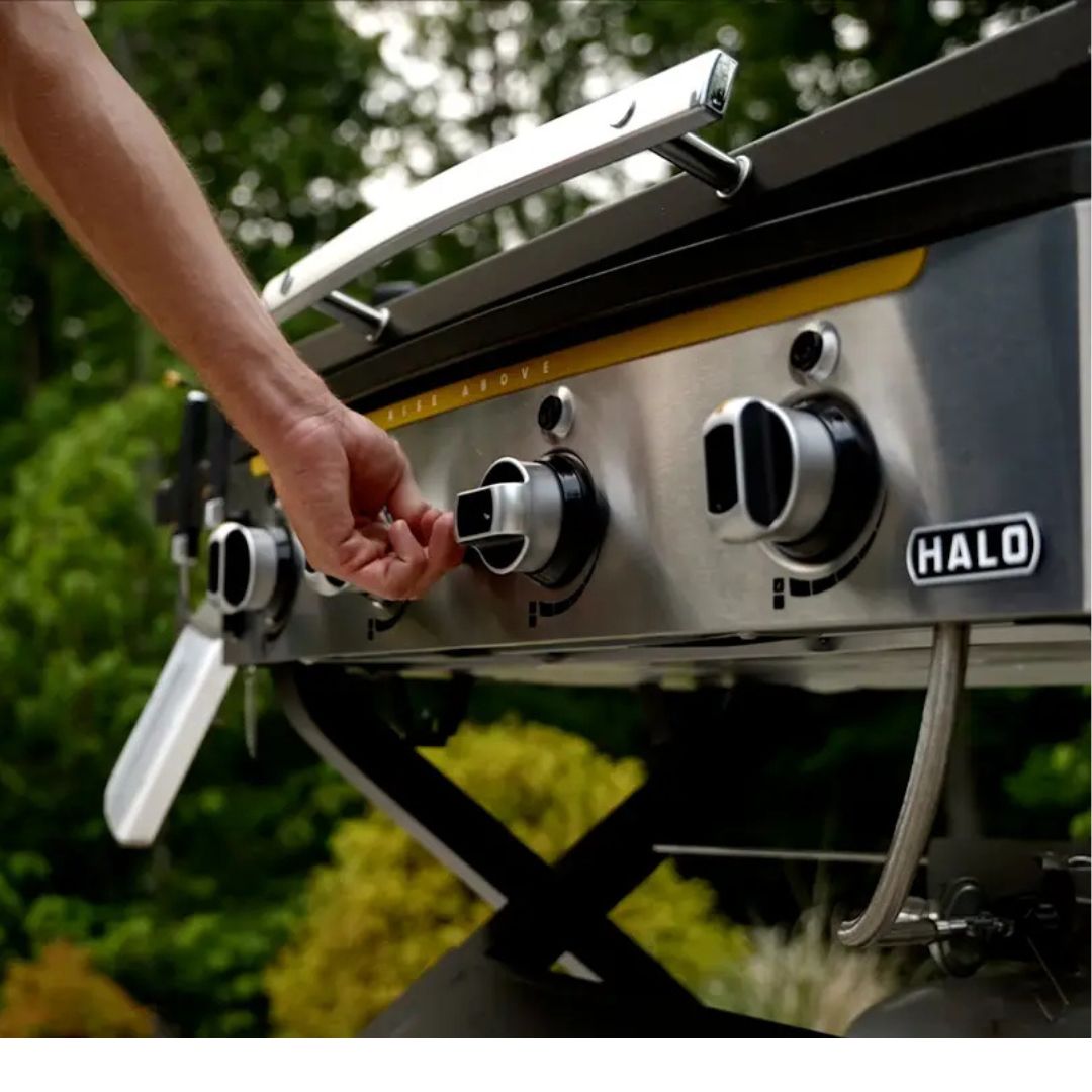 HALO Elite 4B Griddle HZ-1001-XNA 4-burner flat-top stainless steel front with control knobs, hand adjusting a knob, propane hose, outdoor setting