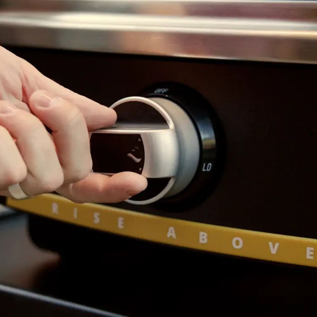 Hand adjusting the silver burner control knob on a HALO Versa 16 outdoor gas pizza oven, black front panel marked "LO" with yellow "RISE ABOVE" trim