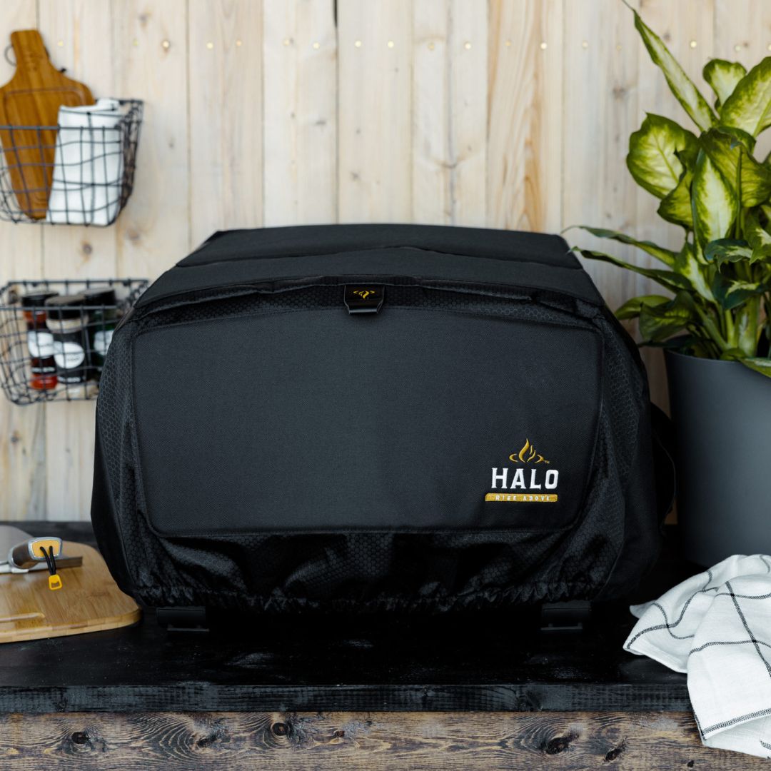 HALO Versa 16 Pizza Oven Cover (HZ-5004) — black fitted weather-resistant cover with elastic hem, top tab and HALO logo, displayed on a wooden countertop in a kitchen setting