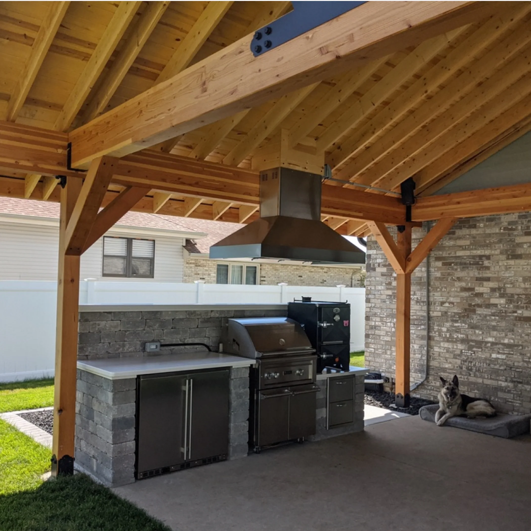 KBU56ASD built-in stainless steel under-counter double-door outdoor refrigerator in gray stone outdoor kitchen island next to stainless gas grill and commercial vent hood under wood-beam pavilion