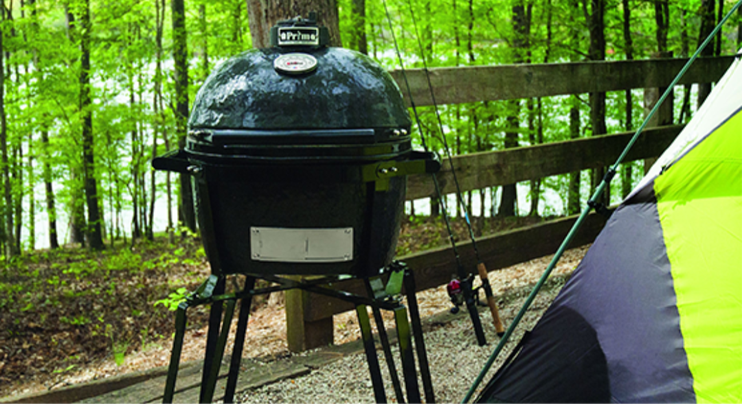 Primo Go portable ceramic kamado charcoal grill on tripod stand with dome thermometer at forest campsite beside tent — portable camping kamado for outdoor charcoal grilling